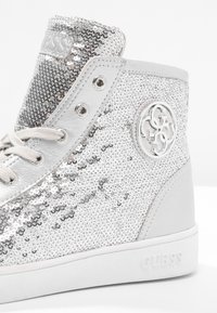 Stříbrné stylové tenisky s flitrovým materiálem a bílými akcenty, obsahující kulaté logo, šněrování v přední části a texturovaný povrch.