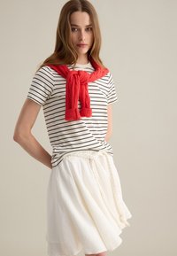 Camiseta de rayas en blanco y negro con mangas cortas, combinada con una falda blanca texturizada y un suéter rojo de punto atado al cuello.