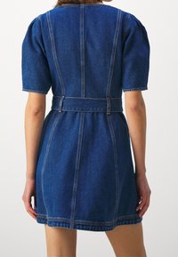Denim-mekko, jossa on lyhyet puff-hihat, vyötäröllä oleva vyö, keskivartalon pituus ja kontrastiväriset tikkaukset. Tumansininen väri, jossa on sileä pinta.