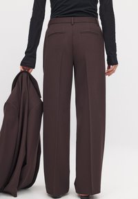 Braune weit geschnittene Hose mit Gesäßtaschen, glatter Textur, flachem Bund und Gürtelschlaufen, kombiniert mit einem passenden Blazer, den man in einer Hand hält.