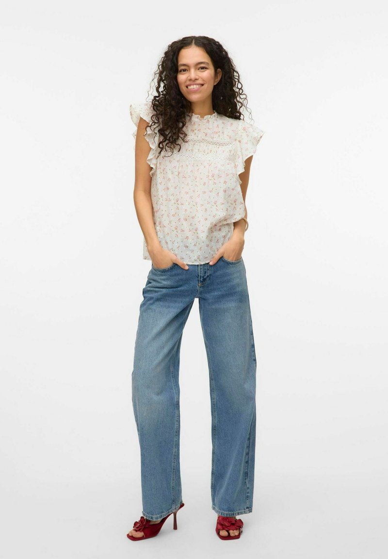Hvid blomster-bluse med flæseærmer, parret med højtaljede lyseblå brede jeans og røde hæle sandals.