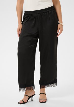 Donna che indossa pantaloni neri a gamba larga con bordo in pizzo all'orlo, sandali neri con tacco e un top bianco, in piedi davanti a uno sfondo neutro.