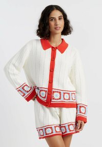 Witte gebreide trui met rode kraag en knopen, met kleurrijke haakdetails op de manchetten en de zoom, gecombineerd met bijpassende shorts.