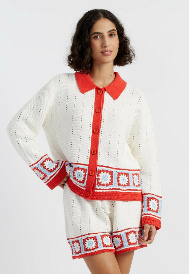 Witte gebreide trui met rode kraag en knopen, met kleurrijke haakdetails op de manchetten en de zoom, gecombineerd met bijpassende shorts.