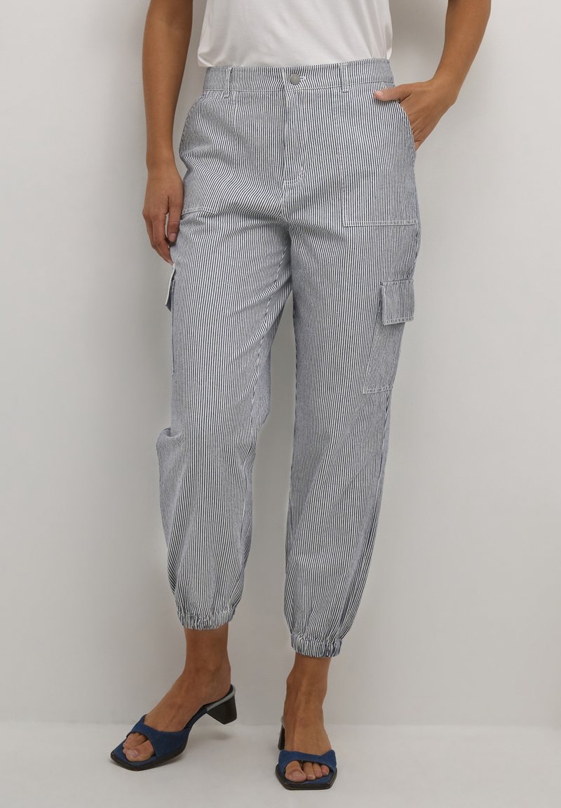 Femme portant un pantalon cargo blanc et bleu à fines rayures, taille haute, longueur cheville, avec des poignets élastiques et des sandales noires à talons ouverts.