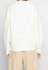 Pull en tricot blanc avec texture côtelée, épaules tombantes et coupe courte. Marquage minimal visible sur le côté gauche.
