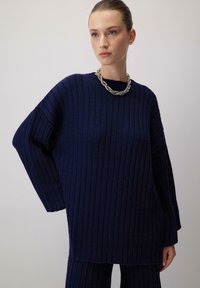 Tumed sinine kudumiga sviiter vertikaalsete soontega, laiade varrukatega ja lõdva lõikega. Koos massiivse hõbedase kaelakeega.