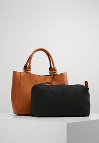 Brązowa skórzana torba tote z podwójnymi uchwytami obok dużej czarnej prostokątnej torby z tkaniny, obie umieszczone na białej powierzchni na szarym tle.