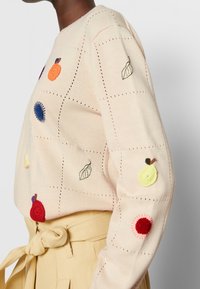 Sweter dzianinowy w jasnoróżowym kolorze z haftowanymi wzorami owoców i liści w różnych kolorach, z dekoracyjnymi dziurkami i okrągłym dekoltem.