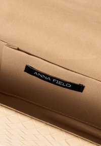Beige stoffen tas met een beige geweven onderste gedeelte. Voorzien van een zwart label met de tekst "ANNA FIELD" in het wit, ter benadrukking van de merkidentificatie.