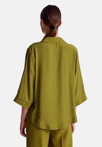 Camicia verde oliva a vestibilità ampia, con una texture morbida, colletto a scendere e maniche tre quarti, caratterizzata da un design pulito e minimalista.