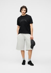 Νεαρός άνδρας με μαύρο μπλουζάκι με σχέδιο, ανοιχτό γκρι σορτς μήκους ως το γόνατο, γκρι κάλτσες, μαύρα αθλητικά παπούτσια, κρατώντας σκούφο σε σκουρόχρωμο φόντο.