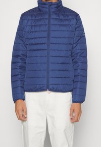 Marineblauwe pufferjas met horizontaal doorgestikt ontwerp, hoge kraag, ritssluiting aan de voorkant en elastische manchetten. Lichtgewicht, gladde stof.