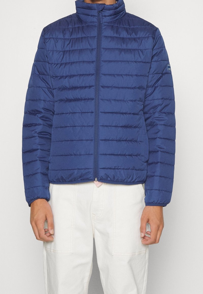 Marineblauwe pufferjas met horizontaal doorgestikt ontwerp, hoge kraag, ritssluiting aan de voorkant en elastische manchetten. Lichtgewicht, gladde stof.
