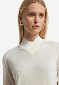 Cremfarbendes Langarmoberteil mit einem strukturierten, texturierten Kragen und einem glatten, leichten Stoff. Das Model trägt große silberne quadratische Ohrringe.