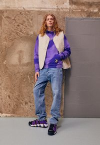 Lila Batik-Hoodie kombiniert mit einer beigen gesteppten Weste, hellblauen Jeans und schwarzen Sneakers mit lila Akzenten und weißen Sohlen.