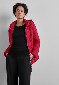 Rote gepolsterte Jacke mit Kapuze, ausgestattet mit einem Reißverschluss und gestepptem Muster. Tragen über einem schwarzen Oberteil und kombiniert mit schwarzen Hosen.