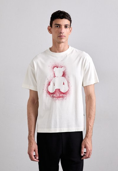 T-shirt blanc en coton avec un graphique d'ours rouge et des accents de splatter. Manches courtes, coupe décontractée et col rond. Pantalons noirs portés en dessous.