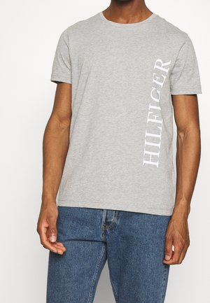 Mężczyzna w jasnoszarej krótkiej koszulce z pionowym napisem „HILFIGER” w białym kolorze po prawej stronie, w połączeniu z niebieskimi dżinsami.