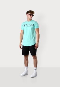 T-shirt à manches courtes bleu clair avec le mot "ESTEEM" en noir, associé à un short noir et des baskets blanches, porté par un homme.