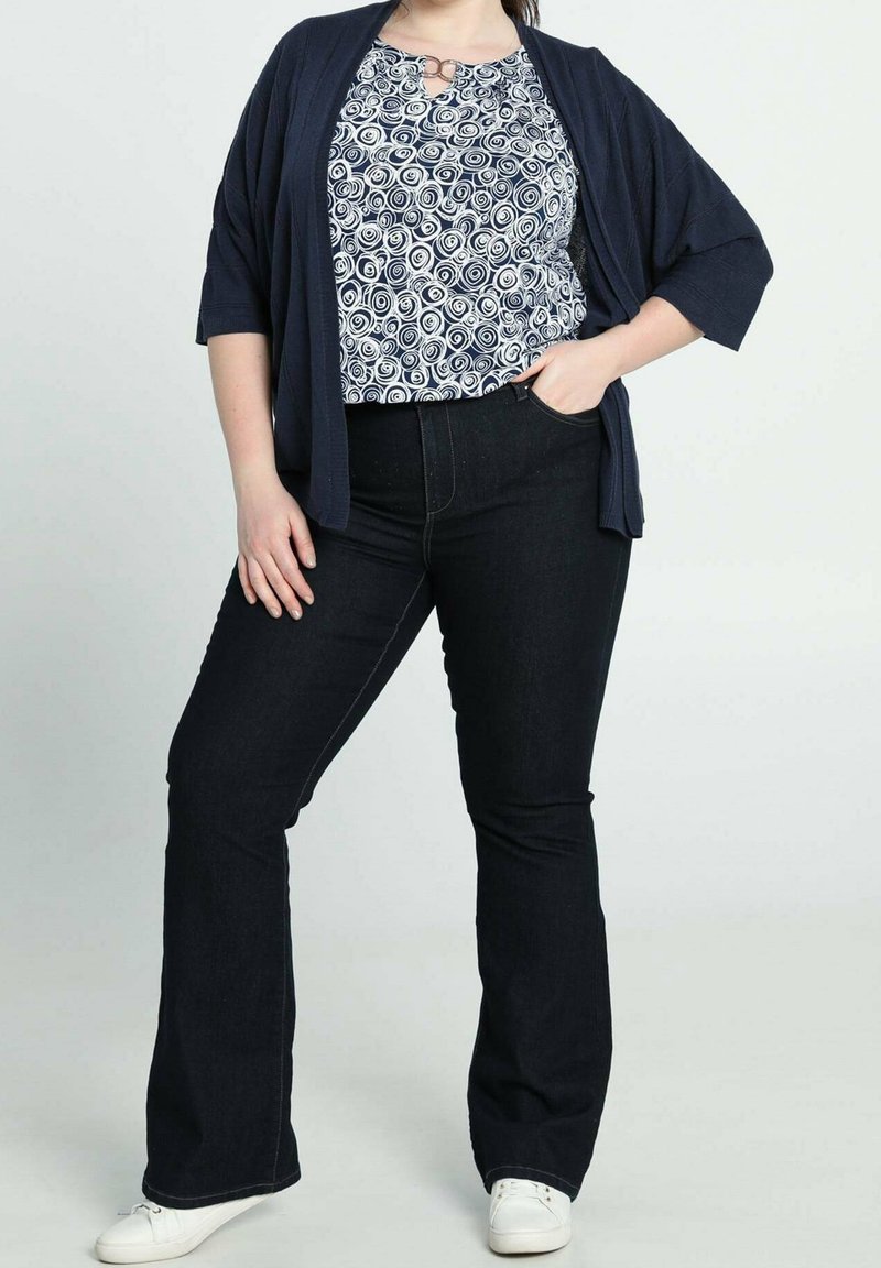 Marinblå cardigan över en vit och marinblå topp med swirl-mönster, i kombination med mörka utsvängda jeans och vita sneakers. Avslappnad och bekväm outfit.