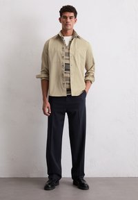 Beige overhemd met knoopjes en een zak, gelaagd over een gebreid patroon. Donkere op maat gemaakte broek en zwarte schoenen maken de outfit compleet.