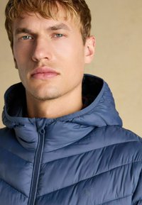 Mørkeblå dunjakke med hætte, prydet med kviltede chevron-mønstre og en frontlynlås. Glat, blank overflade med diskret branding.