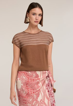 Frau mit kurzen braunen Haaren, die große Blumenohrringe trägt, ein braunes kurzärmliges Oberteil mit gestreiftem oberen Teil und einen roten gemusterten Wickelrock.