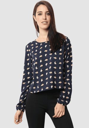 Mujer con blusa de manga larga azul marino con patrones abstractos en beige y blanco, combinada con pantalones negros, de pie contra un fondo liso.