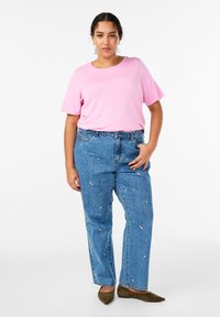 Rozā īsroku T-krekls, kombinēts ar vaļīgām zilām džinsa biksēm ar ziedu izšuvumiem. Modelis arī valkā olīvu zaļas krāsas apavus. Vienkāršs fons.