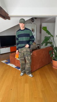 Joven vestido con suéter a rayas, pantalones de camuflaje y gorra, de pie en la sala de estar sosteniendo un cuaderno naranja cerca de un sofá color óxido y una planta grande.