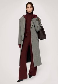 Szary wełniany płaszcz z szerokimi klapami, w zestawie z bordowym golfem i pasującymi spodniami. Modelka trzyma czarną torebkę i ma na sobie bordowe rękawiczki.