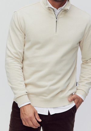 Homme portant un pull à fermeture éclair quart de zip crème sur une chemise blanche et un pantalon marron foncé, une main dans la poche et l'autre détendue.