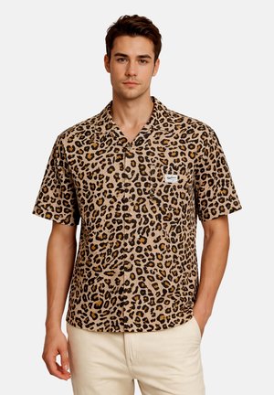 Giovane uomo che indossa una camicia a maniche corte con stampa a leopardo marrone e nero e pantaloni chiari.