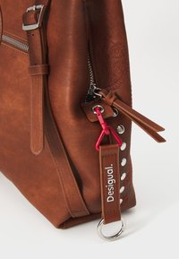 Bruine neplederen handtas met ritssluiting, decoratieve zilveren studs en een rode sleutelhanger. Heeft een gestructureerd oppervlak en dubbele handvatten.