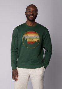 Groene sweatshirt met een cirkelvormige afbeelding van een zonsondergang met horizontale strepen, bergen en silhouetten van bomen en vogels.