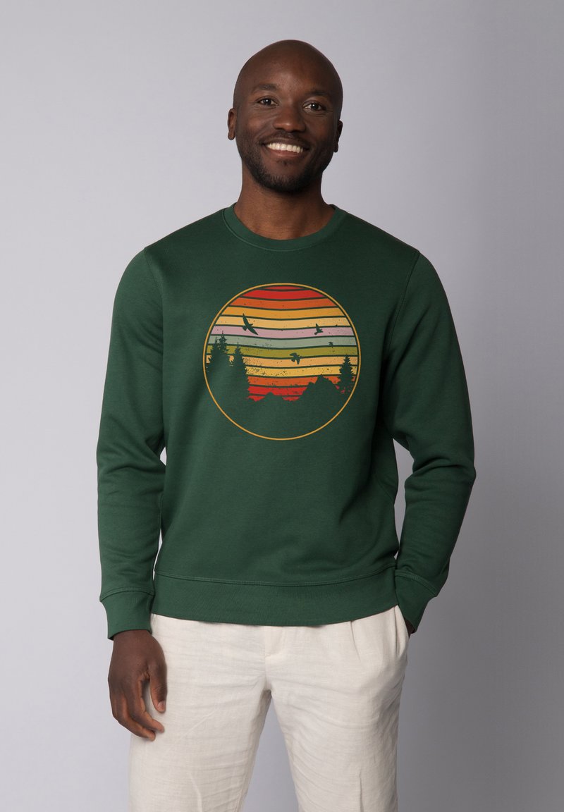 Groene sweatshirt met een cirkelvormige afbeelding van een zonsondergang met horizontale strepen, bergen en silhouetten van bomen en vogels.