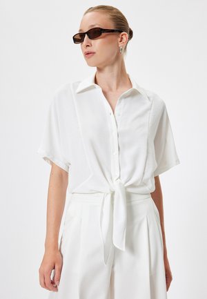 White short-sleeve blouse with a collar and front tie detail. Soft fabric with a smooth texture. Worn with high-waisted white trousers.
