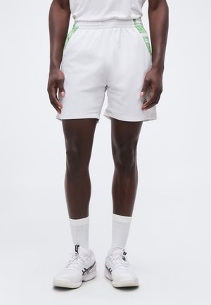 Sportlane, kes kannab valgeid spordipükse roheliste detalidega, valgeid keskmise pikkusega sokke ja valgeid jooksujalatseid, seisab tavalise tausta ees.