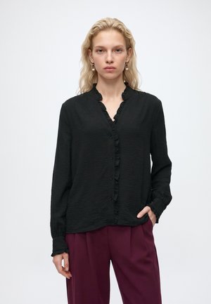 Jeune femme aux cheveux blonds portant un chemisier noir à manches longues avec des volants et un pantalon violet foncé, debout avec une main dans la poche.