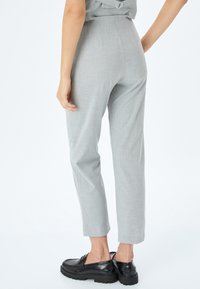 Grau zugeschnittene Hose aus strukturiertem Stoff, schmale Passform und knöchellanges Design. Getragen mit schwarzen Slippern mit einer klobigen Sohle.