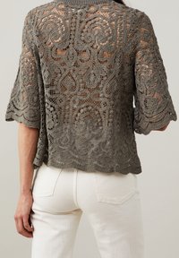 Haut en crochet gris présentant des motifs en dentelle complexes, manches courtes, et un ourlet festonné, associé à un pantalon blanc, mettant en valeur le design à maille ouverte.