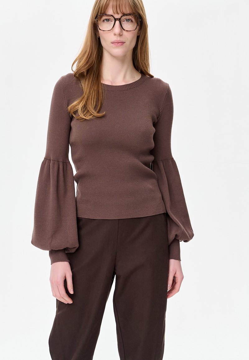 Vrouw met lang rood haar en bril, gekleed in een bruine geribde trui met ballonmouwen en bijpassende bruine broek, naar voren gericht.