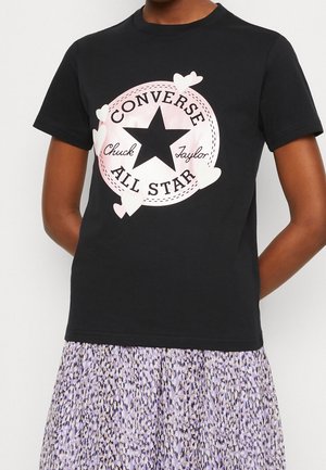 Osoba nosząca czarną koszulkę Converse All Star z różowymi sercami i fioletową spódnicę w wzory.