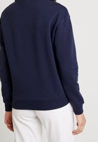 Tmavě modrá mikina s žebrovanými manžetami a lemem, jednoduchým vzorem a měkkou texturou, nošená přes bílé kalhoty.