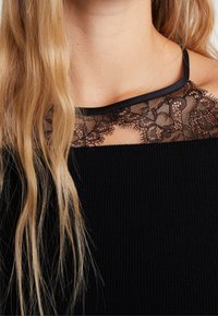 Haut noir à texture côtelée, avec un empiècement en dentelle à motifs floraux et une bordure en satin le long de l'encolure.