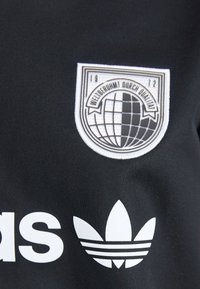 Melnas sporta apģērbs ar izteiktu balto Adidas logo un apļveida izšuvumu, kurā ir globuss un teksts vairoga formā.