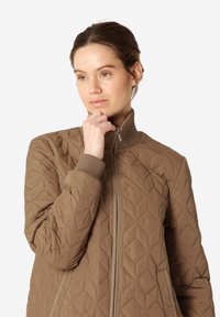 Chaqueta acolchada marrón con cuello alto, con un patrón de diamantes, cierre de cremallera y puños acanalados. Textura suave y diseño cálido.