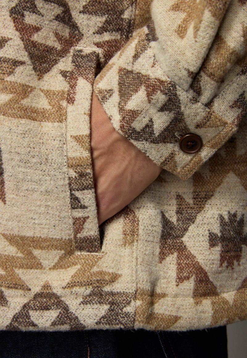 Main dans la poche d'une veste beige et marron à motifs avec poignet boutonné et design géométrique.