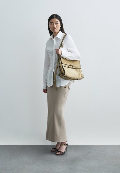 Γυναίκα φορώντας λευκό μπλούζα, μπεζ μακριά φούστα, μαύρες γόβες ανοιχτού ποδιού, κρατώντας μια μεγάλη μπεζ τσάντα ώμου, στέκεται σε γκρι δάπεδο μπροστά σε άσπρο τοίχο.